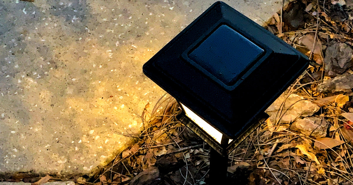 A solar light sits at the corner of a cement path and shines during the night. Twigs, leaves, and rocks surround the light.