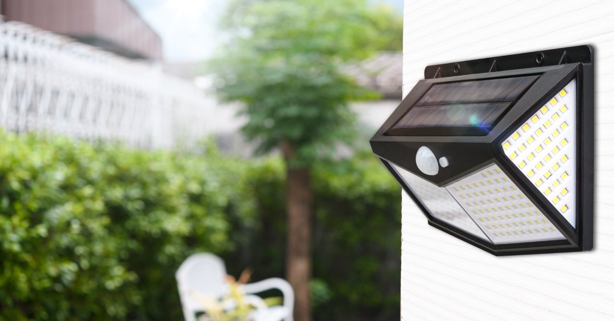 A wall-mounted solar light soaks up energy during the daytime hours. A home's backyard is in the background.